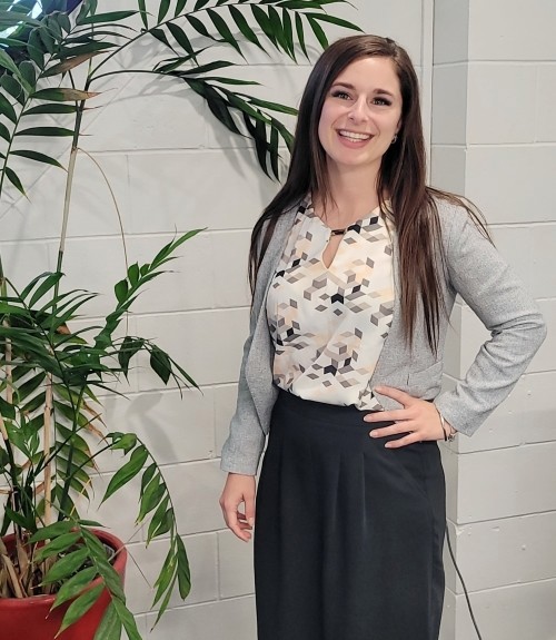 Sharleena Wilson, Relationship Manager at Safety 'n Action, smiling and standing in front of a white wall and indoor plant, wearing a patterned blouse and black skirt.