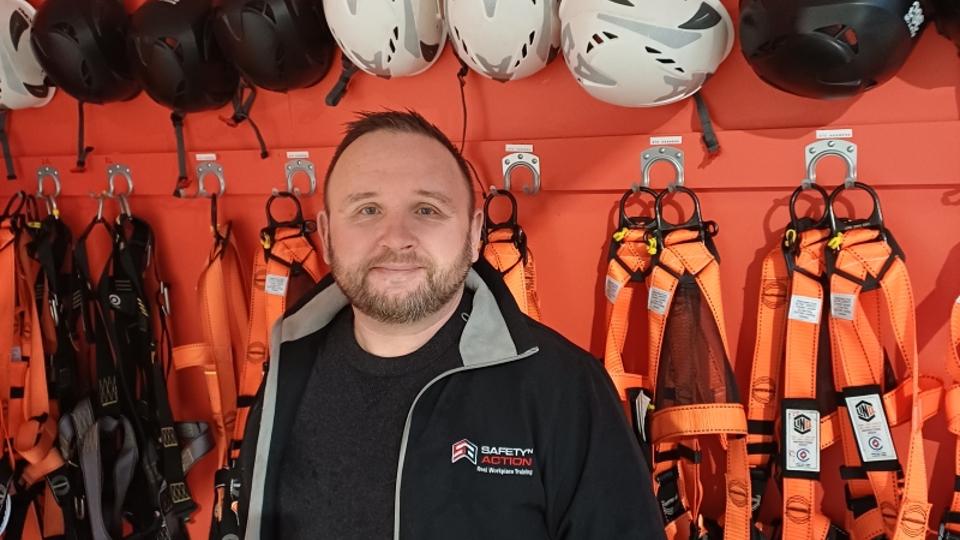 Christopher Petitpas smiling in front of the height safety harness and helmet wall at the Safety 'n Action Tauranga training centre.
