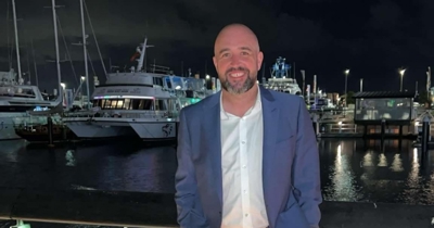 Gary Lowis smiling at the Auckland Viaduct, dressed in a blue suit with boats lit up in the marina behind him.