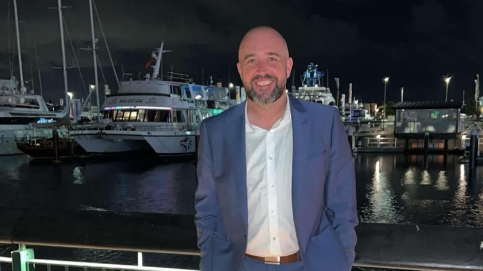 Gary Lowis smiling at the Auckland Viaduct, dressed in a blue suit with boats lit up in the marina behind him.