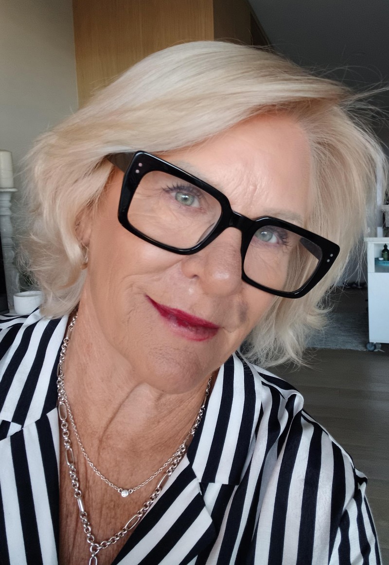Sheryl Jones smiling in a striped shirt and black-framed glasses, with blonde hair and layered silver necklaces, indoors.