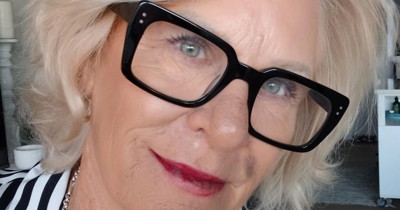 Sheryl Jones smiling in a striped shirt and black-framed glasses, with blonde hair and layered silver necklaces, indoors.
