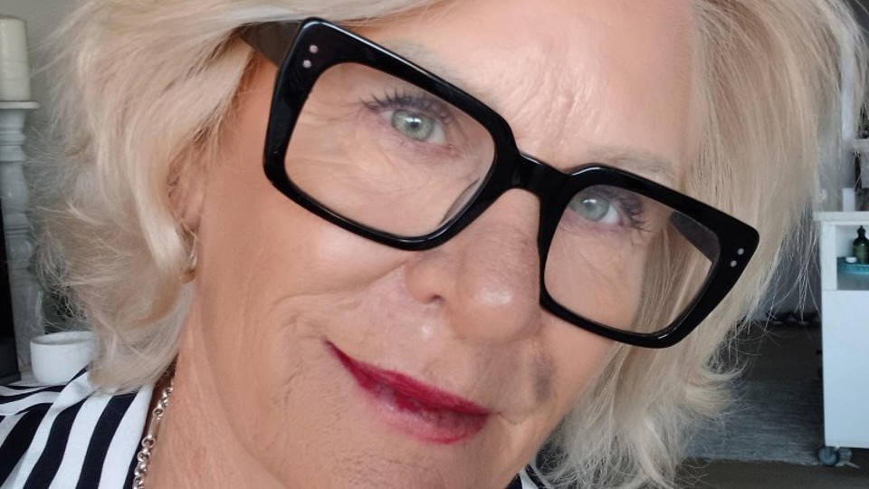 Sheryl Jones smiling in a striped shirt and black-framed glasses, with blonde hair and layered silver necklaces, indoors.